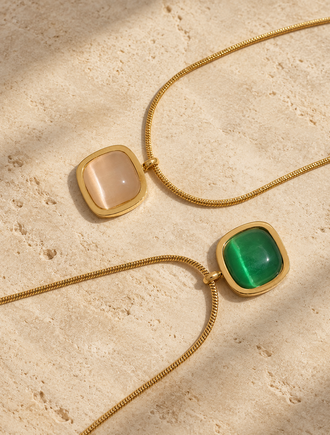 Two gold chain necklaces with square bezel settings, one featuring a white agate and the other a vivid green agate stone, displayed on a textured beige stone surface.