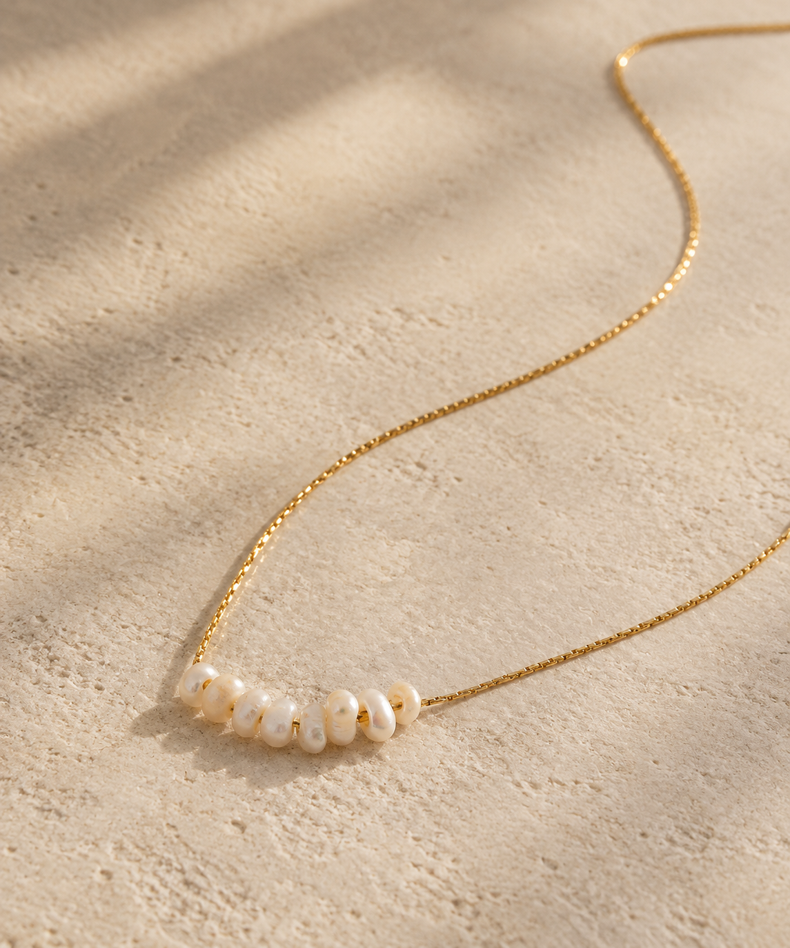 A delicate gold chain necklace featuring a row of small, irregular white shell beads, displayed on a textured beige surface with soft leaf shadows.