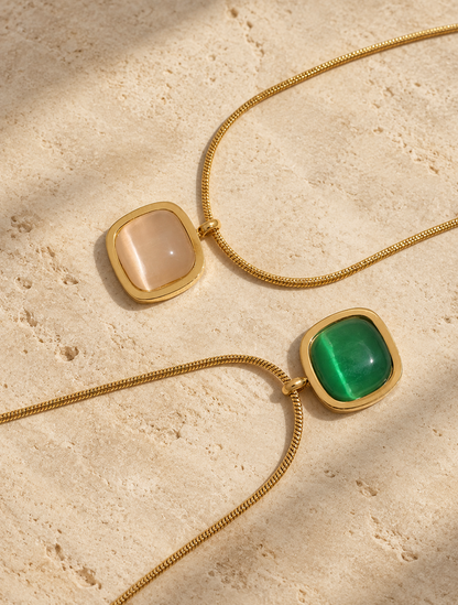 Two gold chain necklaces with square bezel settings, one featuring a white agate and the other a vivid green agate stone, displayed on a textured beige stone surface.