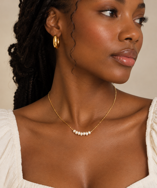 A close-up of a woman wearing a delicate gold chain necklace with a row of small white fresh water pearls 